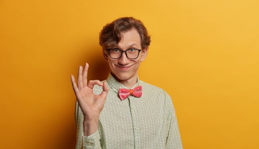 A young man with glasses and a bow tie showing the ok sign on a yellow background.