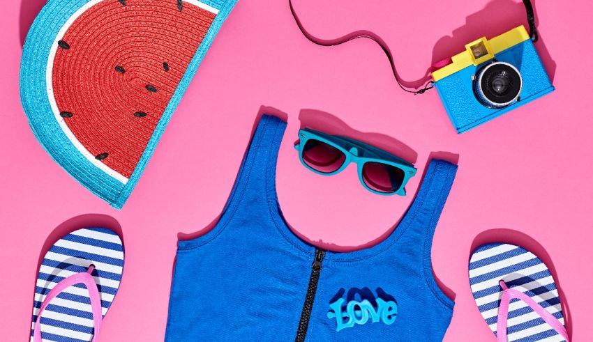 Watermelon, sunglasses, flip flops and a camera on a pink background.