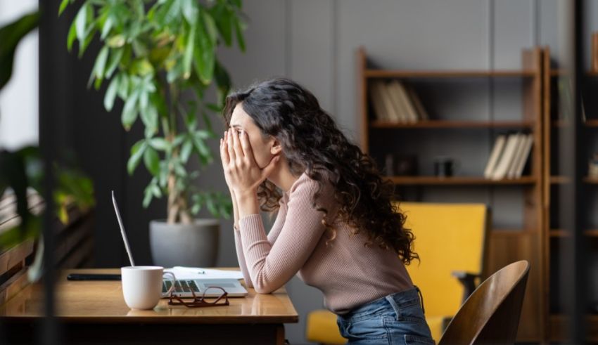 A woman covering her face while sitting at a desk with a laptop.