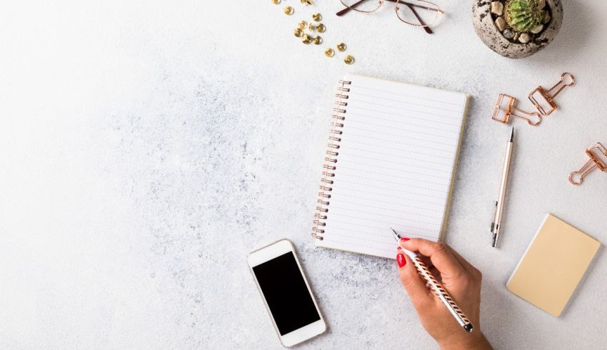A woman's hand writing on a notepad next to a phone and a plant.