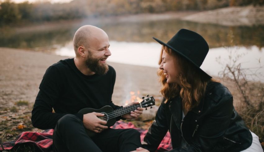 A couple playing ukulele on a blanket near a lake.