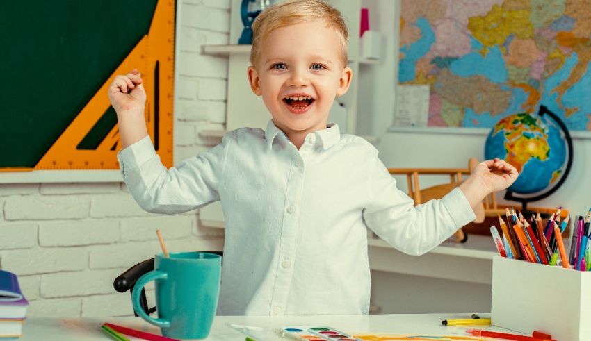 A young boy is standing in front of a desk with a cup of crayons.