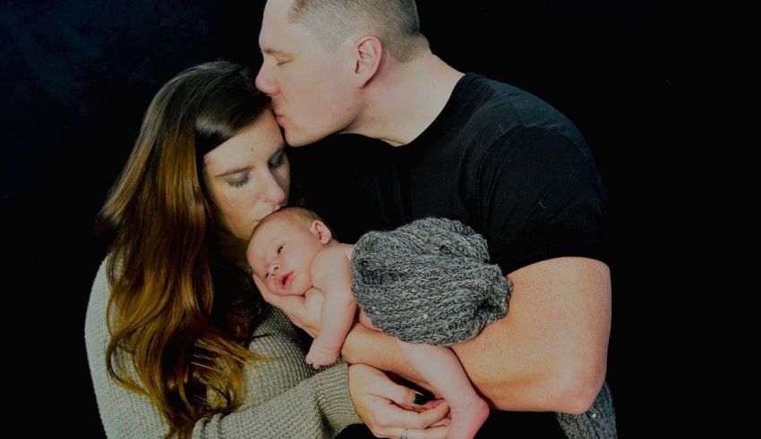A man and woman kissing their newborn baby in front of a black background.