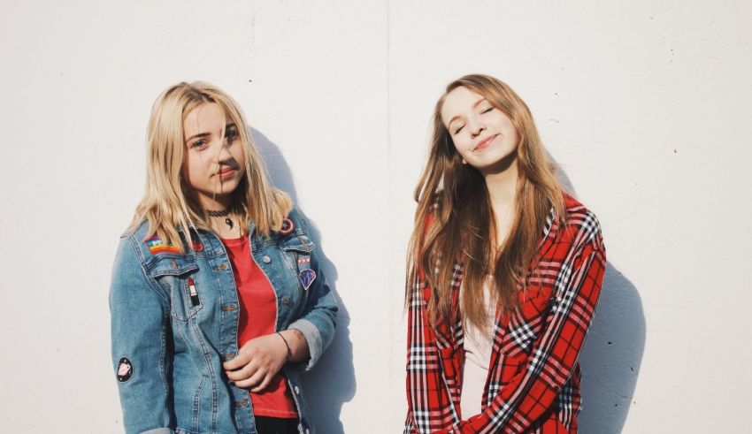 Two young women standing next to each other in front of a white wall.