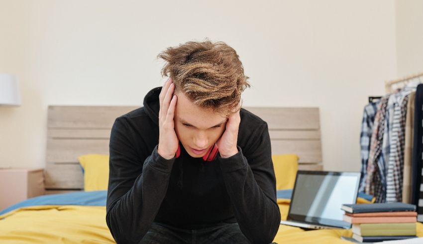 A young man sitting on his bed with his hands on his head.