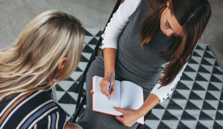 Two women sitting on a chair and writing in a notebook.