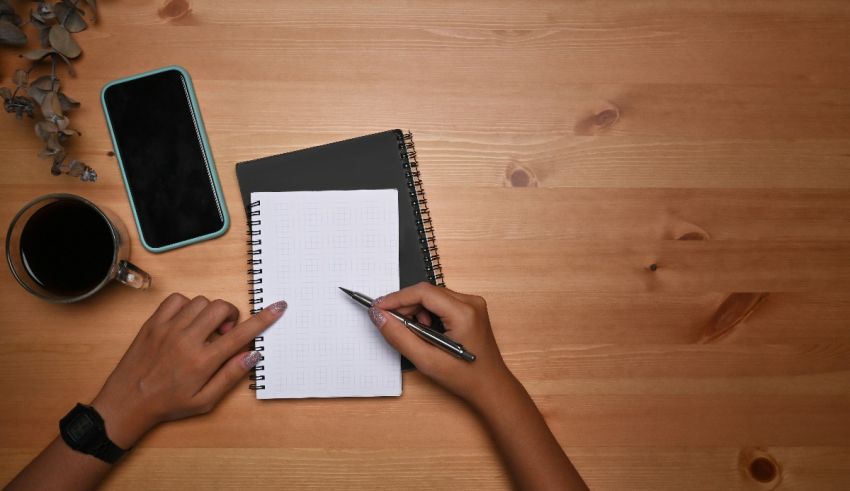 A woman's hand writing on a notebook on a wooden table.