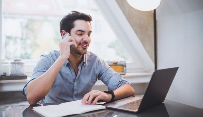A man is talking on the phone while sitting at a desk with a laptop.